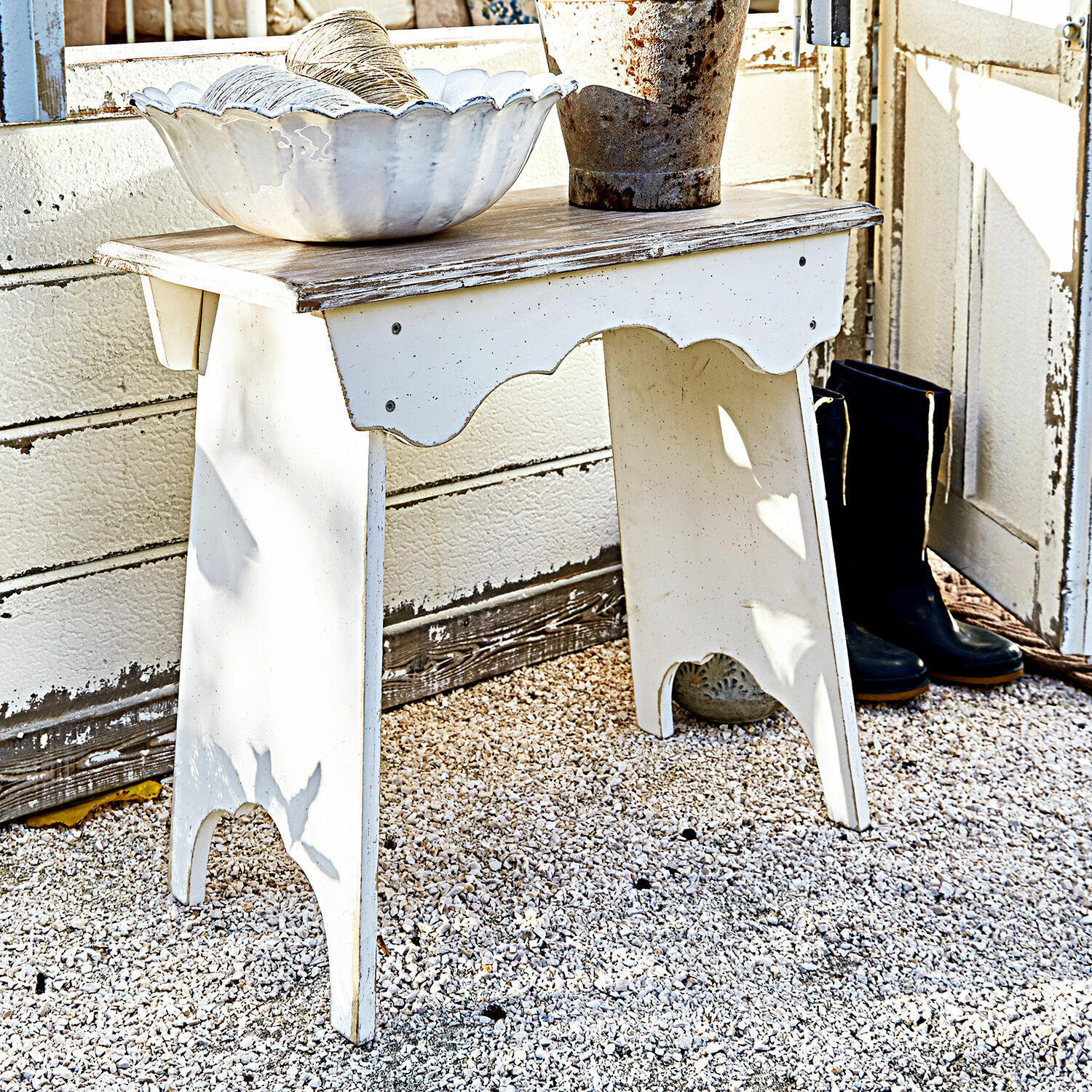 Kruk Fria Witte houten tafel met verweerde afwerking op grindvloer met decoratieve schaal en zwarte laarzen