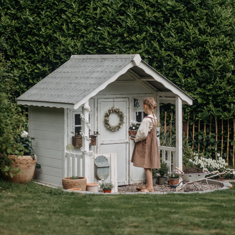 Witte houten tuinhuisje met decoratieve krans en planten, kind houdt bloempot vast