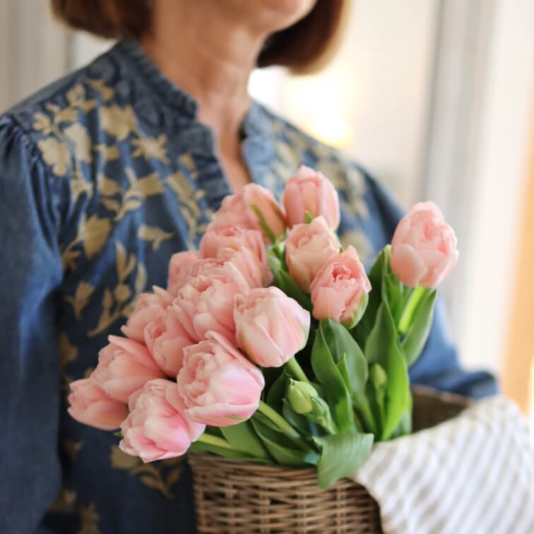Boeket roze tulpen in een rieten mand gehouden door vrouw in blauwe blouse met bloemenpatroon