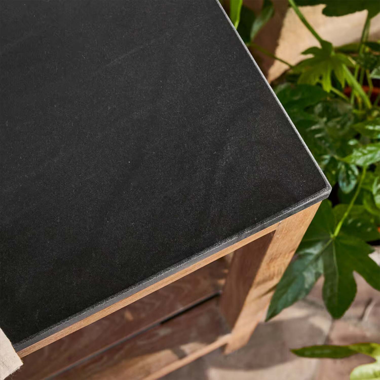 Tafelblad van zwart steen met houten onderstel omringd door groene planten, tuinmeubel