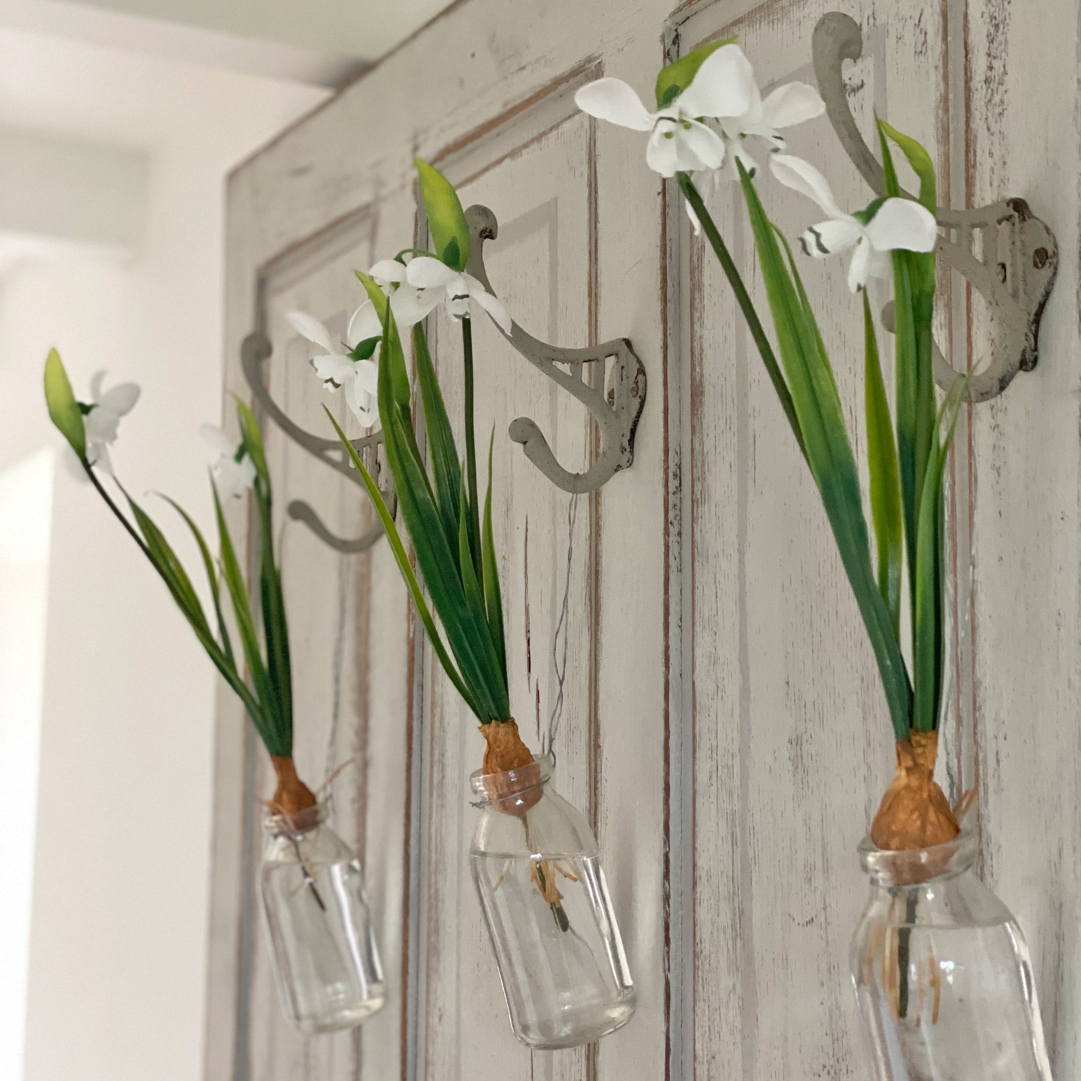Drie witte bloemen in glazen flesjes aan metalen haken aan een houten deur