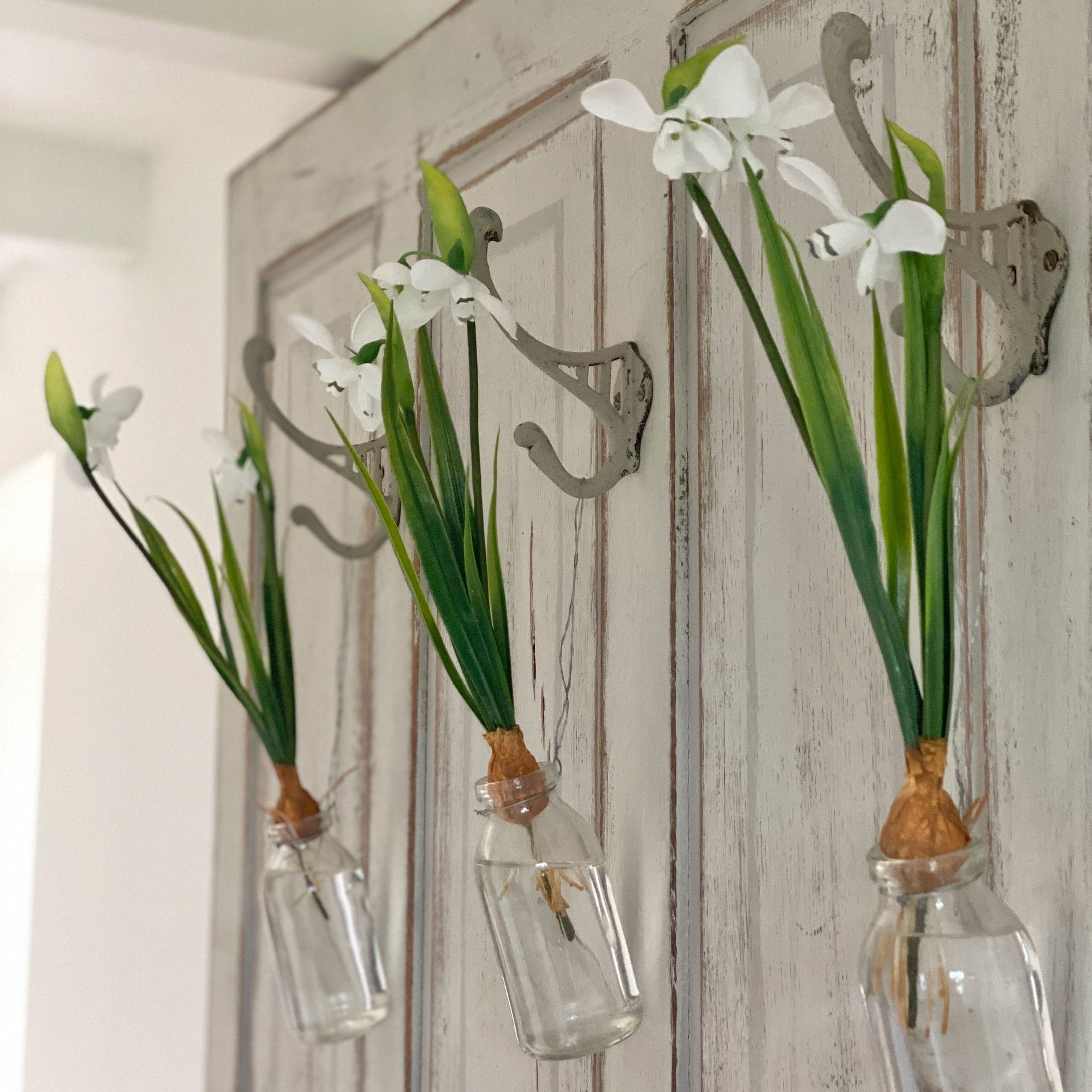 Drie witte bloemen in glazen flesjes aan metalen haken aan een houten deur