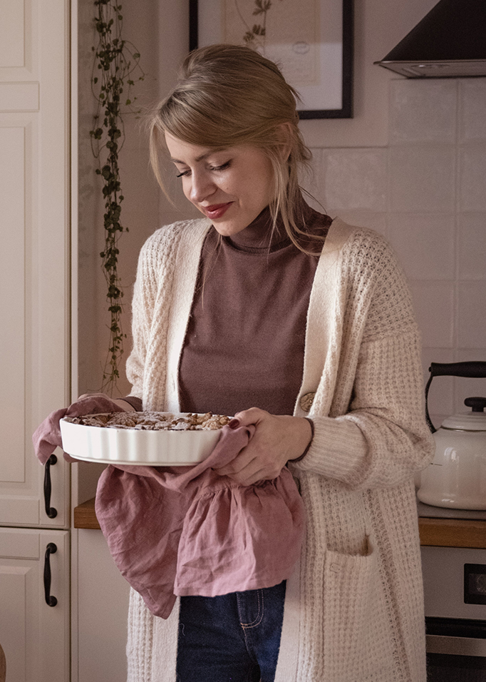 Vrouw in beige gebreid vest houdt witte keramische ovenschaal met roze doek vast in keuken