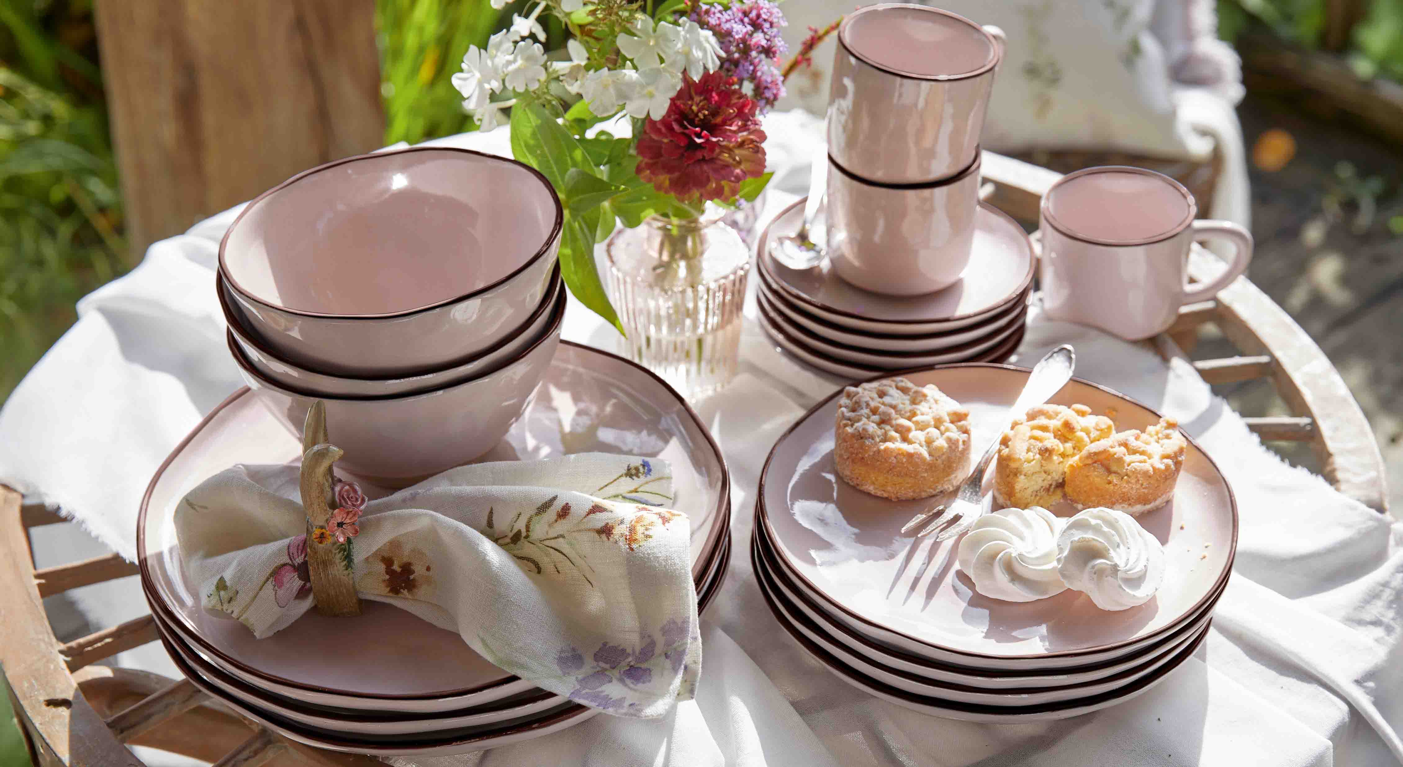 Set roze keramisch servies met borden, kommen en mokken op houten tafel met gebak en bloemen