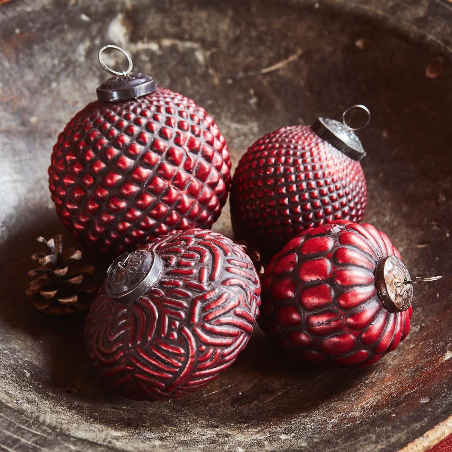 Vier rode kerstballen met verschillende structuren in een houten schaal met dennenappels