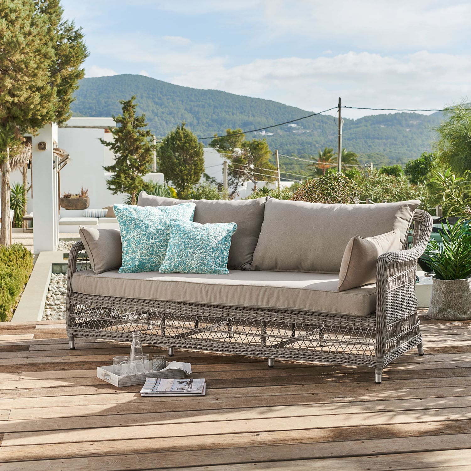 Bank Tanaiste Tuinbank van grijs rattan met beige kussens en turquoise sierkussens op houten terras