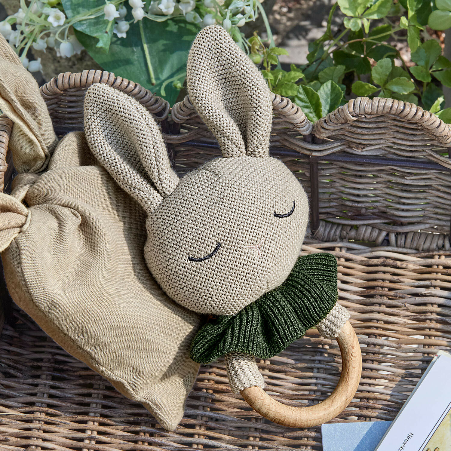 Beige gebreide konijnenrammelaar met groene kraag en houten ring op een rieten achtergrond