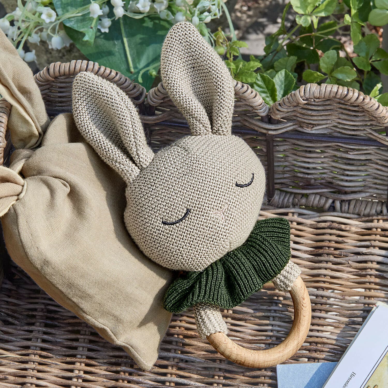 Speelring Cuddlecloud Beige gebreide konijnenrammelaar met groene kraag en houten ring op een rieten achtergrond