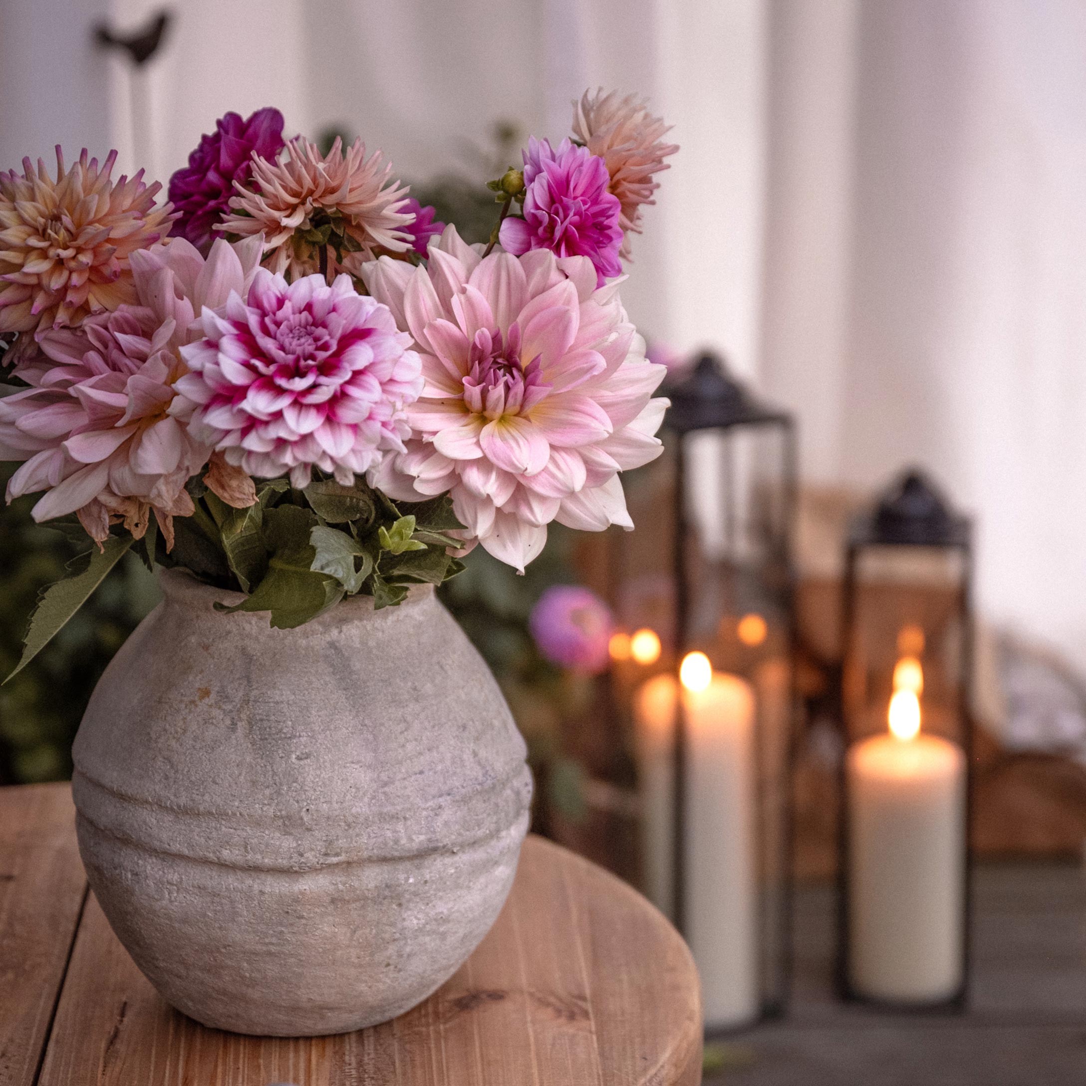 Grijze keramische vaas met roze-paarse bloemen op houten tafel, lantaarns en brandende kaarsen op de achtergrond