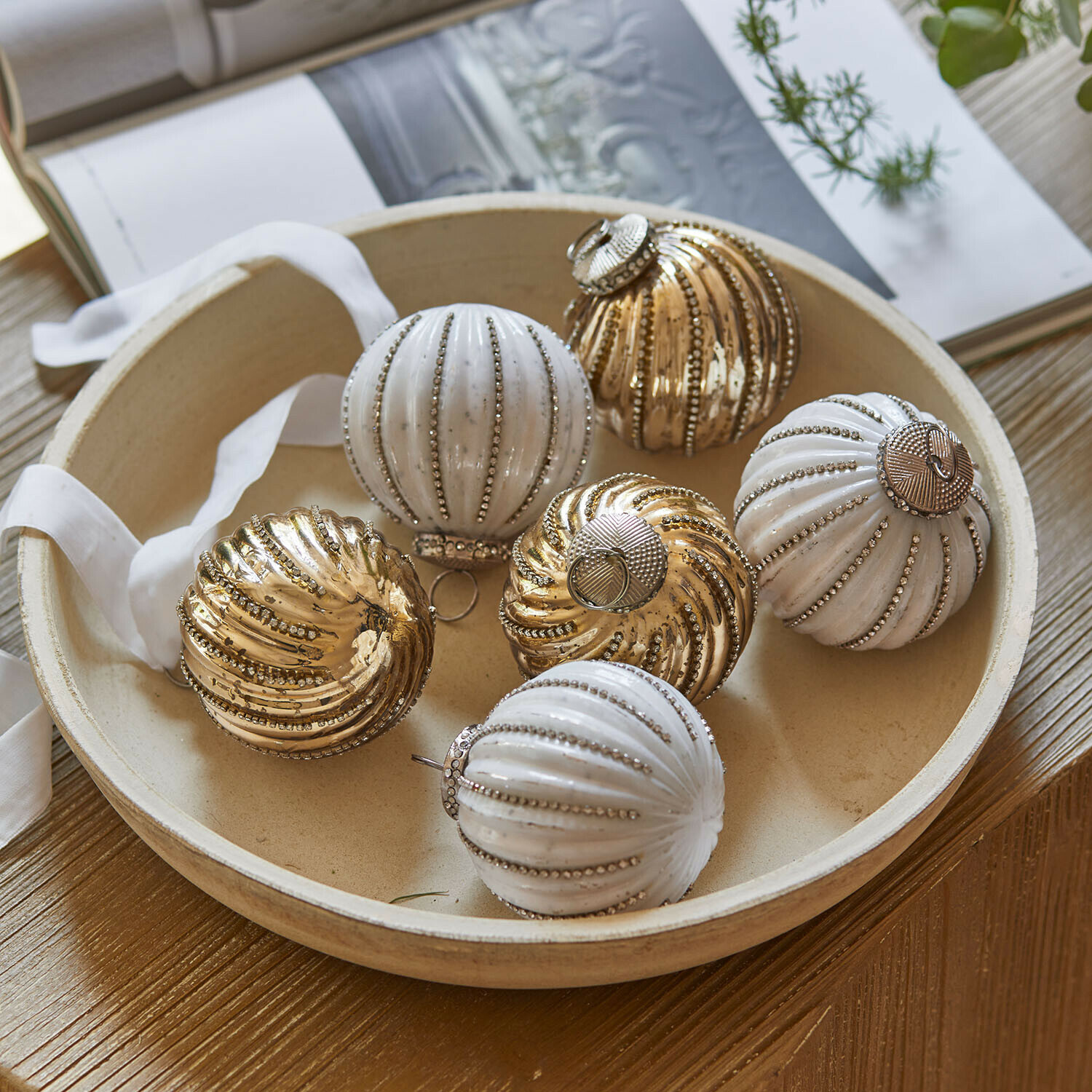 Kerstbal set van 6 Shaela Set witte en gouden kerstballen met metallic glans in een ronde schaal op een houten tafel