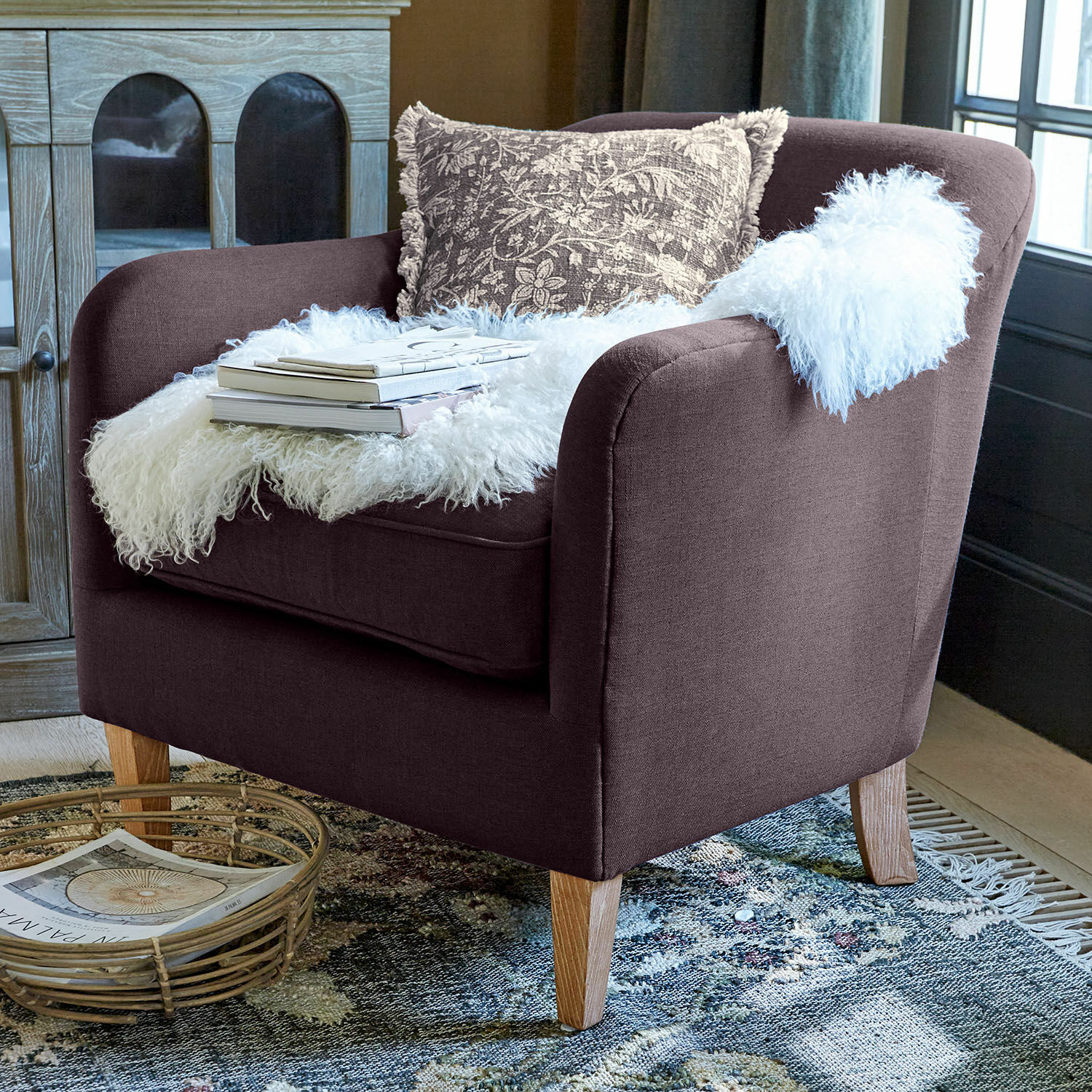 Fauteuil Jefferson Bruine gestoffeerde fauteuil met houten poten, versierd met een witte bontachtige deken en een patroon kussen
