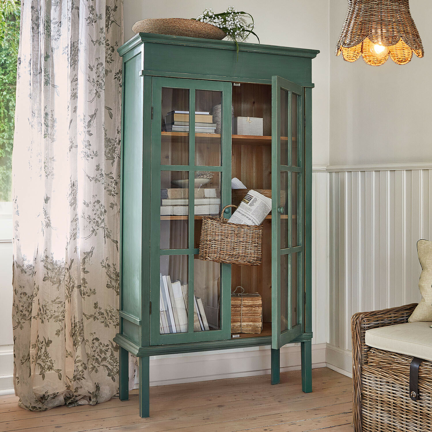Groene houten vitrinekast met glazen deuren en rieten mand in lichte woonkamer met natuurlijk licht