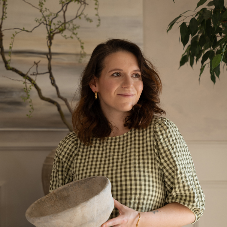 Vrouw in groene geruite jurk houdt grote grijze keramische kom vast in lichte kamer met planten