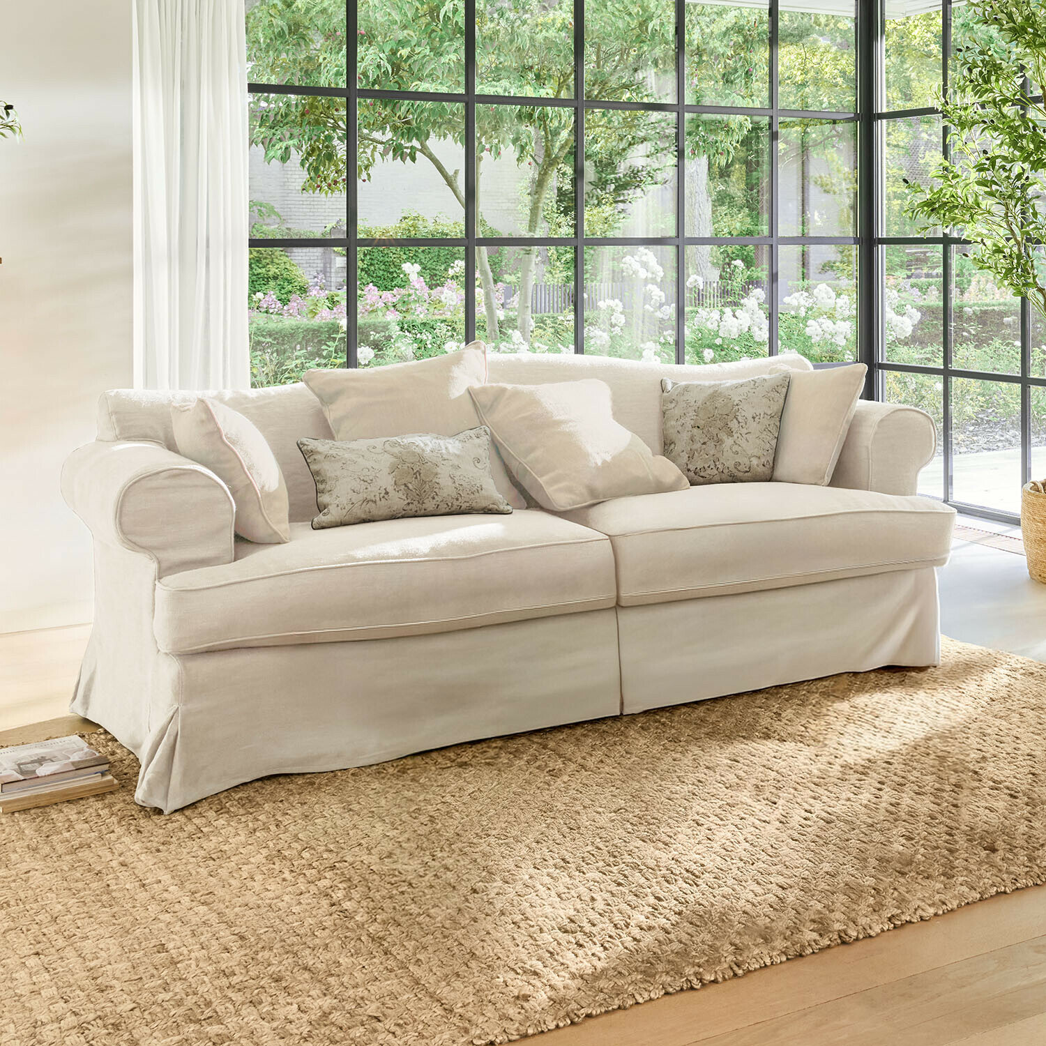 Beige stoffen bank met sierkussens op natuurlijke vloerkleed in lichte woonkamer met grote ramen