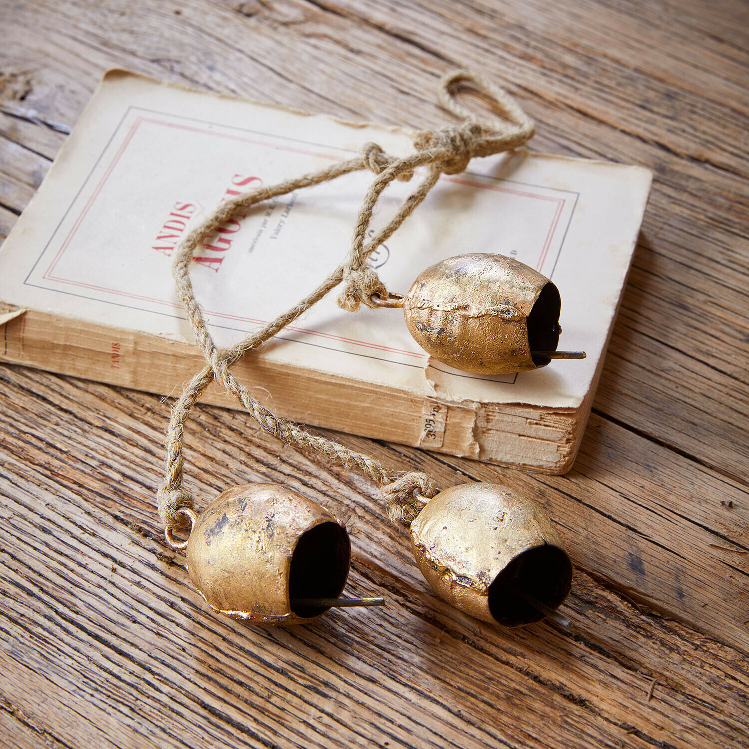 Bel set van 3 Orelli Drie gouden metalen bellen aan een touw op een houten tafel met een oud boek erachter