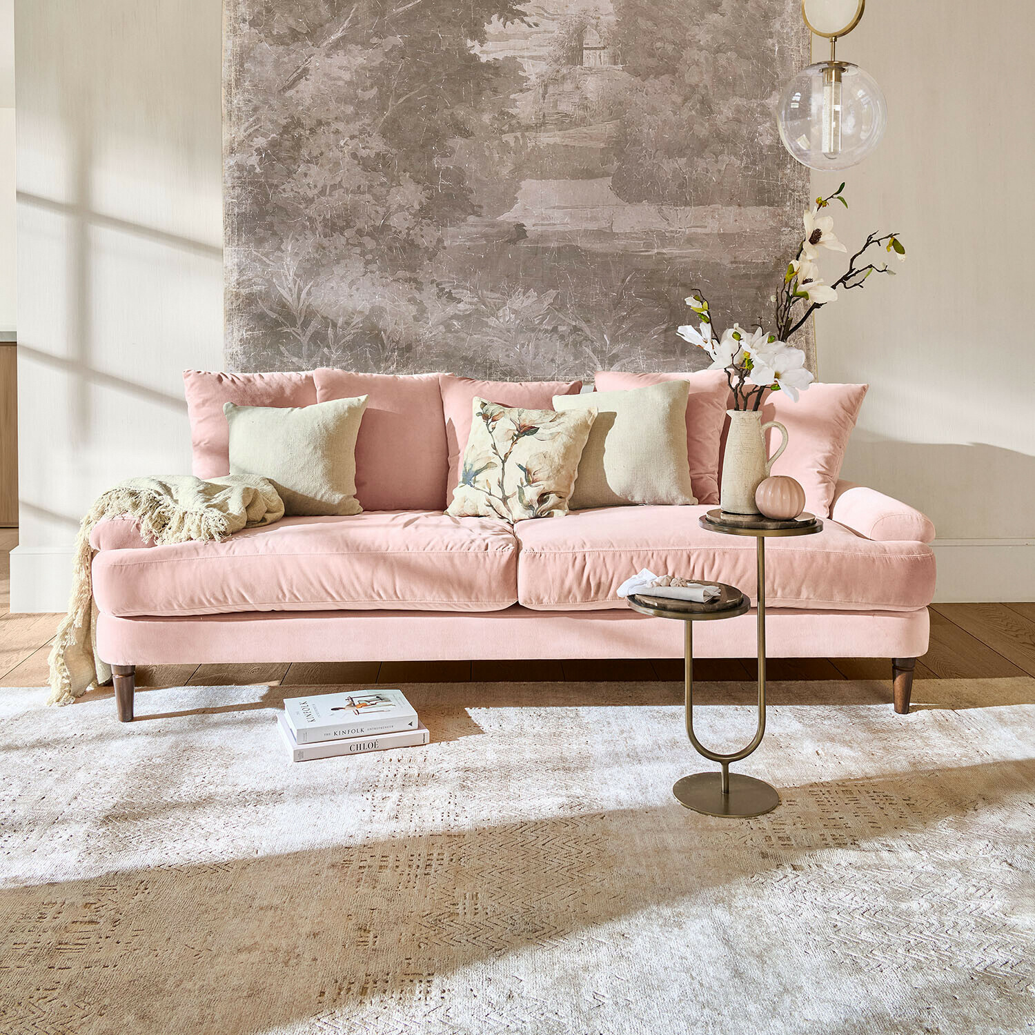 Roze fluwelen bank met kussens en bijzettafel in lichte woonkamer met wanddecoratie