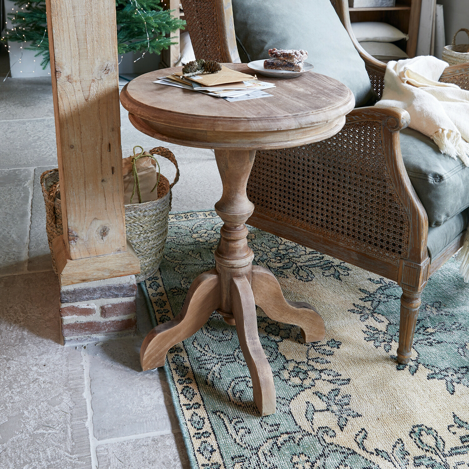 Bijzettafeltje Mengomeyen Ronde houten bijzettafel in natuurlijke kleur naast een rotan stoel op een patroon vloerkleed