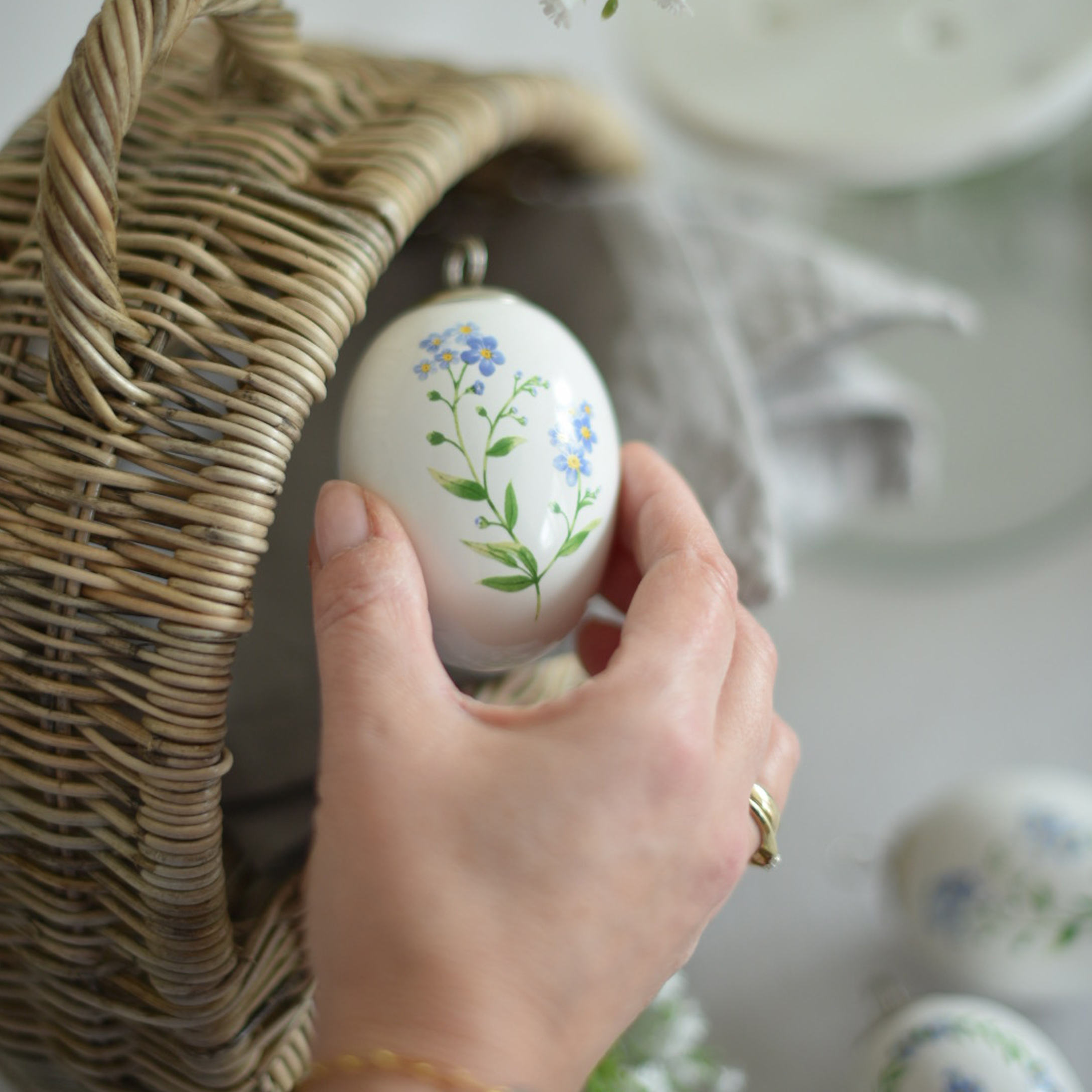 Hand houdt wit keramisch paasornament met blauwe bloemen naast een rieten mand