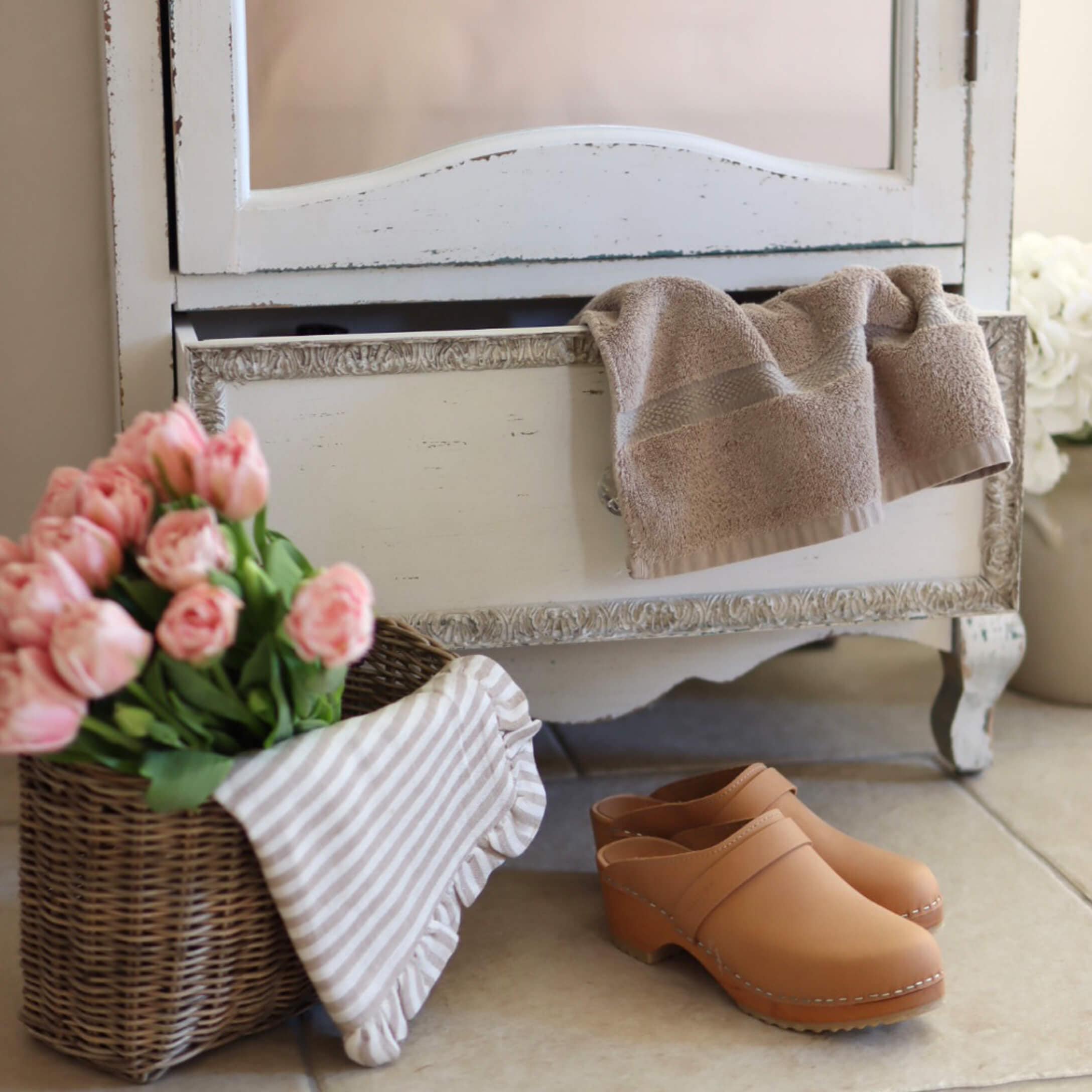 Beige leren klompen op de vloer naast een rieten mand met roze bloemen en een handdoek