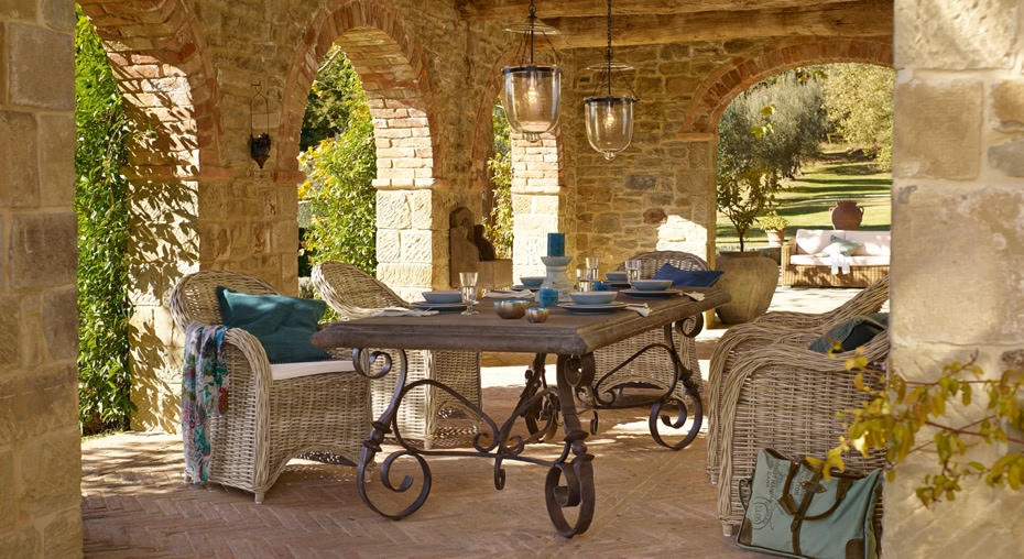 Stijlvolle tafel met smeedijzeren poten en rotan stoelen onder een stenen boog op terras