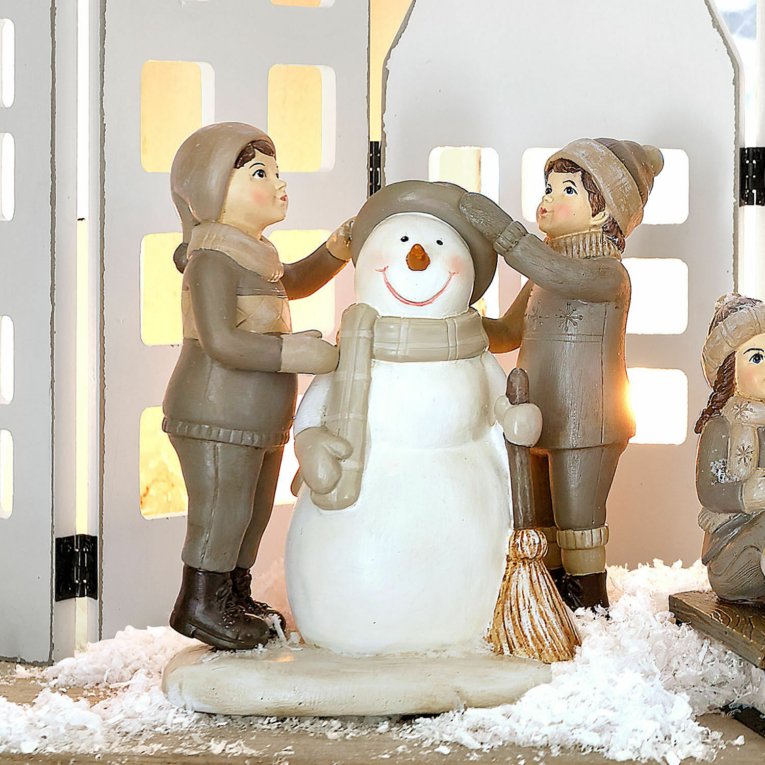 Sierbeeldje Elvirette Beelden van kinderen die een sneeuwpop van materiaal in beige en bruine tinten aankleden op decoratief sneeuw