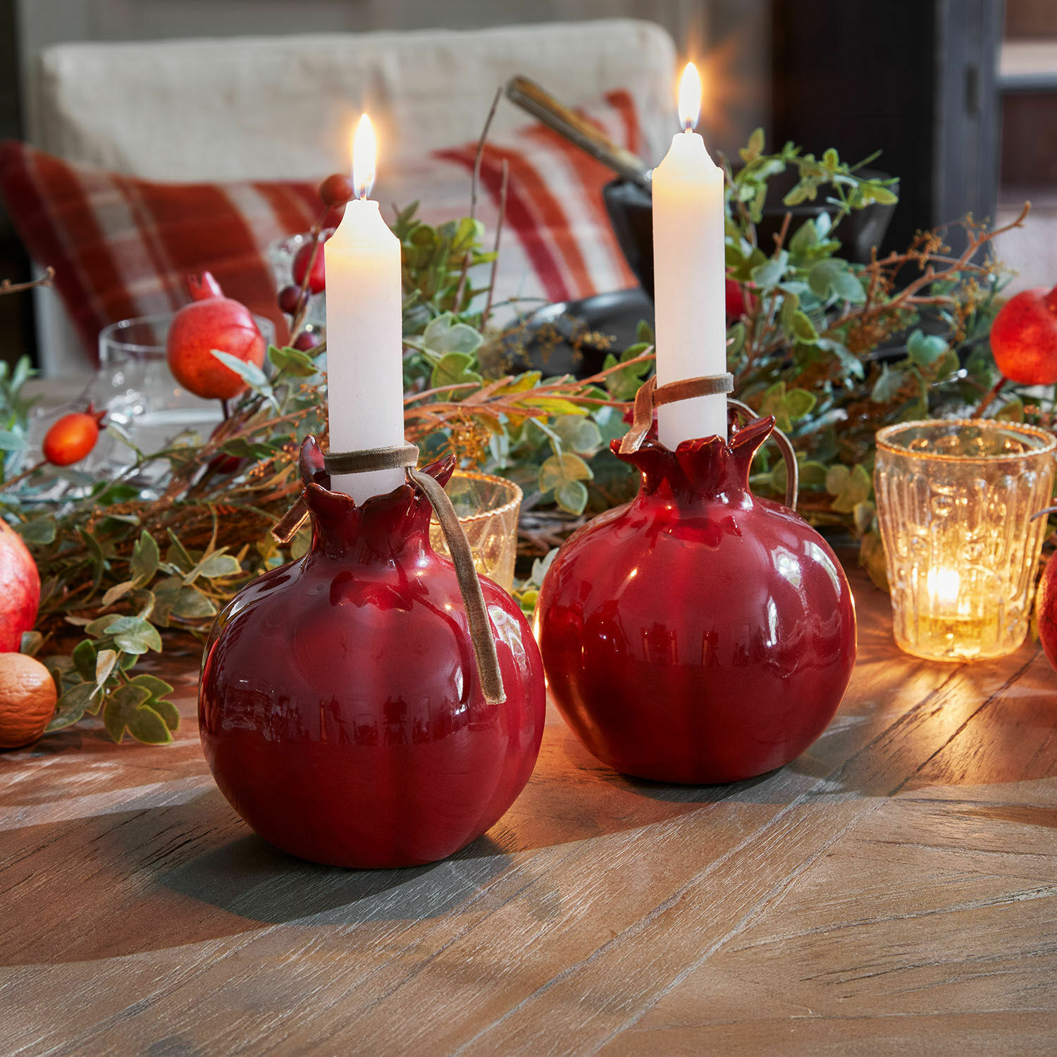 Twee rode keramische kandelaren in granaatappelvorm met witte kaarsen op een houten tafel met decoratie