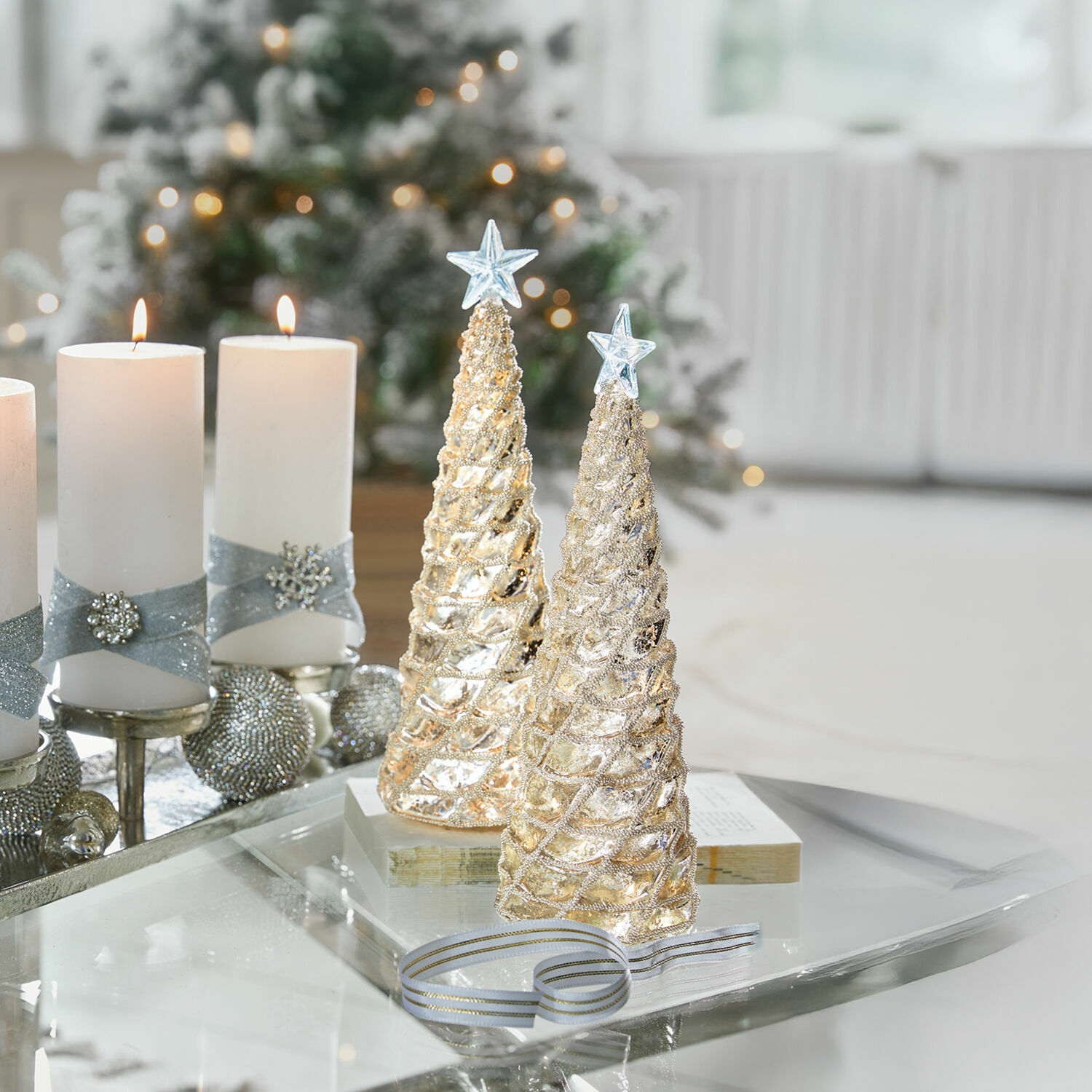 Twee gouden kerstboomdecoraties met sterren bovenop op glazen tafel met witte kaarsen en feestelijke achtergrond