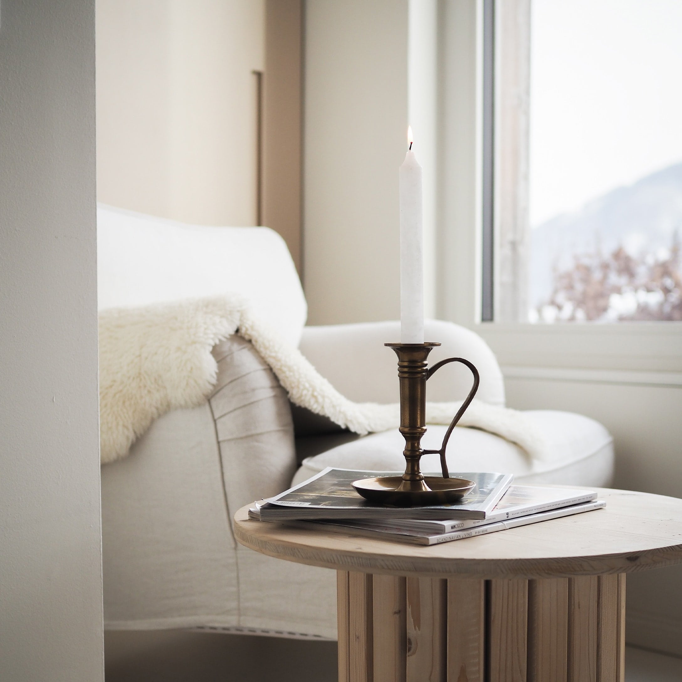 Messing kandelaar met witte kaars op houten tafel naast witte bank en deken