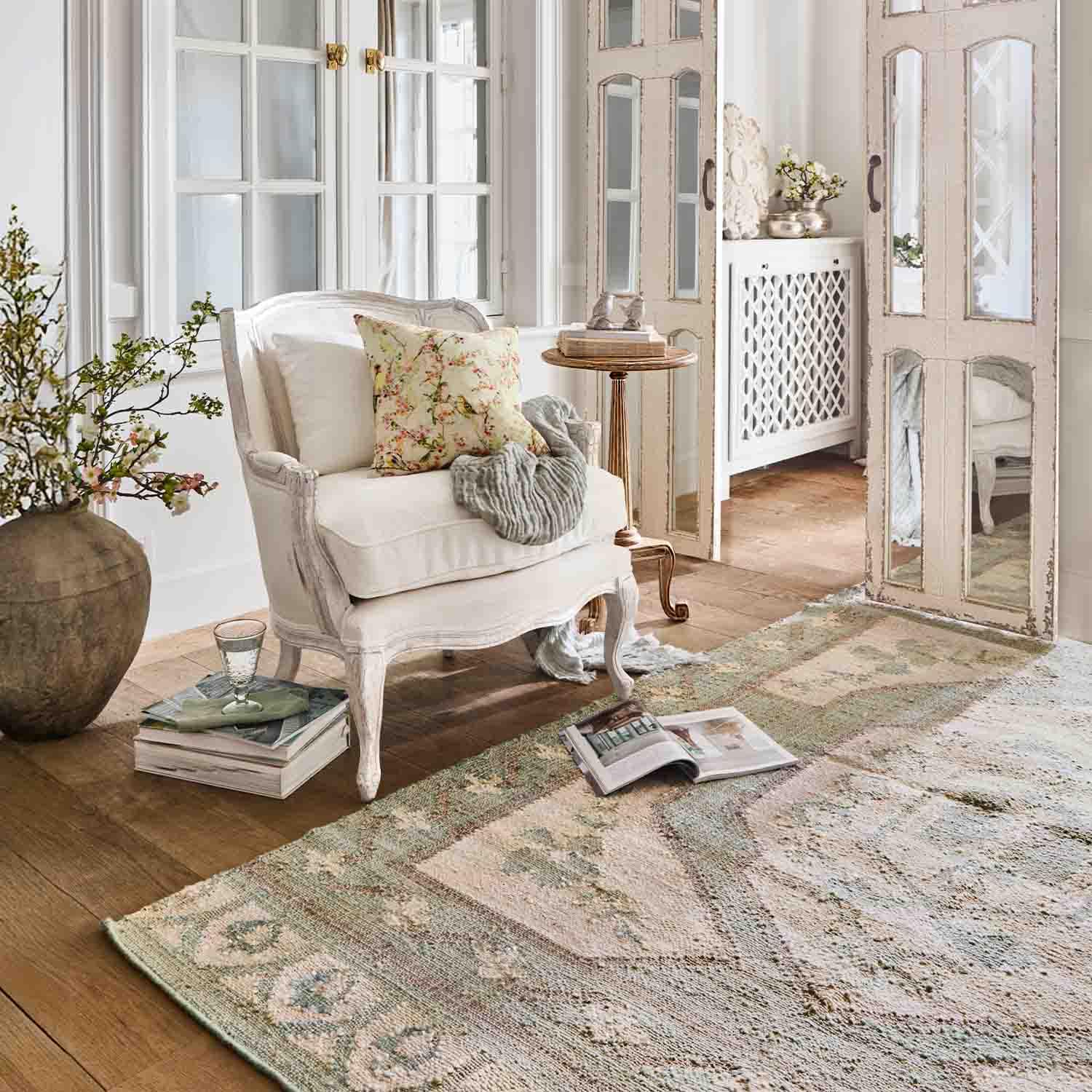 Witte houten fauteuil met bloemenkussen in lichte woonkamer met vloerkleed en decoratieve spiegel